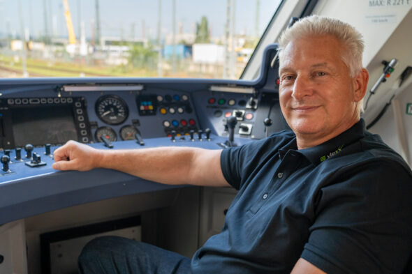 Ein erfahrener Triebfahrzeugführer mit grauen Haaren und schwarzem Poloshirt sitzt entspannt im Führerstand eines modernen Zuges. Er lächelt und blickt in die Kamera, während seine Hand auf dem Bedienpult mit zahlreichen Knöpfen, Hebeln und Anzeigen ruht. Durch das Fenster sind Bahnanlagen und Industriegebäude im Hintergrund zu sehen.