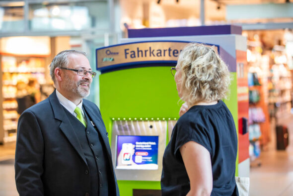 Ein Fahrgastbetreuer und eine Frau unterhalten sich vor einem grünen vlexx-Fahrkartenautomaten.