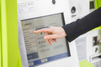 Eine Person zeigt auf das Display eines Fahrkartenautomaten von vlexx.